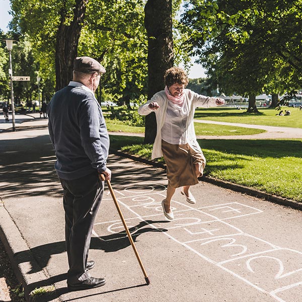 Seniorin springt wie junges Mädchen durch Himmel und Hölle Spiel im Park und ihr Mann schaut zu