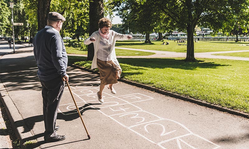 Seniorin springt wie junges Mädchen durch Himmel und Hölle Spiel im Park und ihr Mann schaut zu